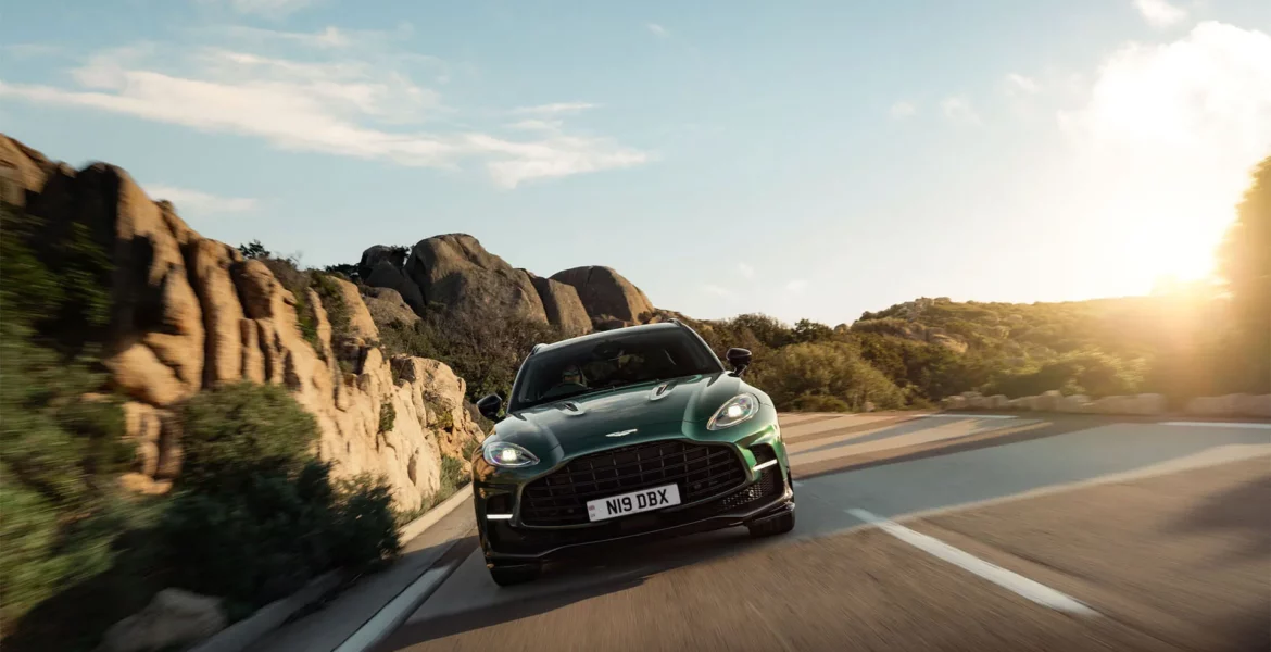 An Aston Martin car driving on a tarred mountain road with the sun in the background sky