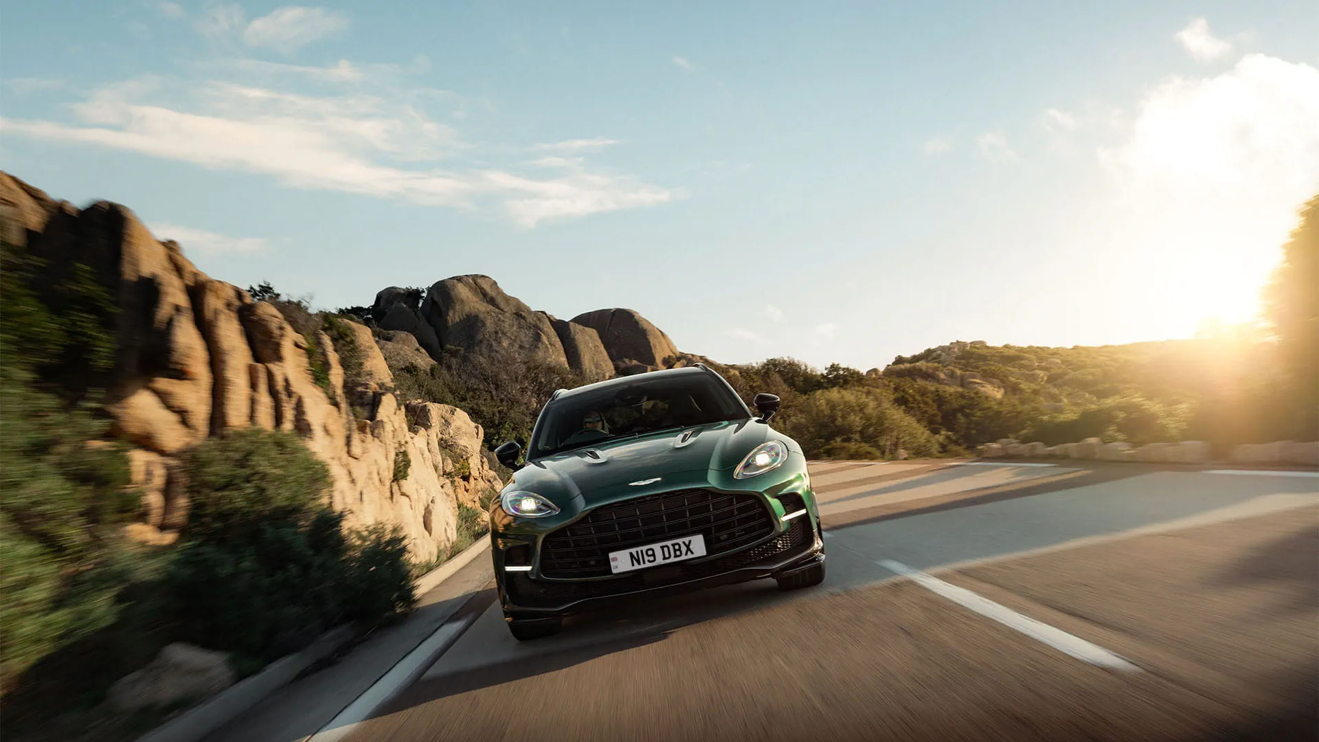An Aston Martin car driving on a tarred mountain road with the sun in the background sky